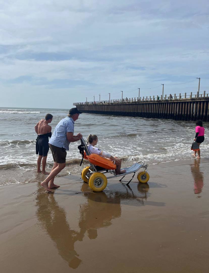 Sue Martin using an all-terrain beach wheelchair on Scottburgh Beach with the ocean behind her.